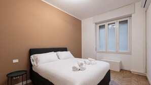 A bedroom with a bed, a small table, a window, and a wall-mounted air conditioner. - CORSO GARIBALDI 64 - LIVING MILAN (Milan)