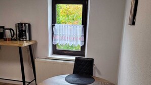 A dining area with a round table, two black chairs, a window with a view of greenery, a radiator, and a coffee maker on a wooden table. - Fitters' apartment for two  (Schleiden)