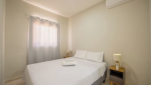 Un lit bien fait, avec du linge de lit blanc, une table de chevet avec une lampe et un climatiseur fixé au mur.