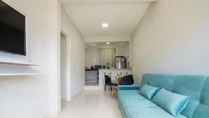 Un salon moderne avec un canapé de couleur turquoise, un téléviseur à écran plat fixé au mur et un coin cuisine à l’arrière-plan.