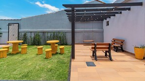 Un espace patio avec des tables et des bancs en bois, une pergola illuminée de guirlandes lumineuses et un petit jardin agrémenté de plantes en pot.
