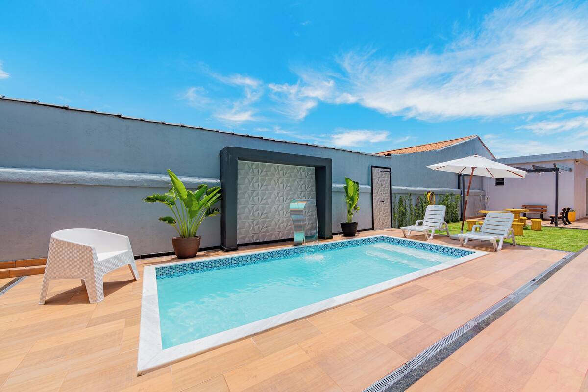A swimming pool with a tiled deck, a white chair, a potted plant, and a patio area with a table and chairs.