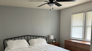 Ein Schlafzimmer mit einem Bett, einem Nachttisch, einer Kommode, einem Deckenventilator und einem Fenster mit Jalousien.