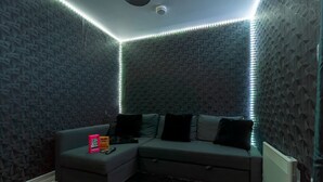 A corner room with a grey sectional sofa, a black rug, and LED strip lighting along the ceiling. - Emerald Suite- Bham Citycentre- Luxury-cinema Room (Birmingham)