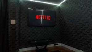 A room with a wall-mounted TV displaying the Netflix logo, a black entertainment table, and a patterned wall. - Emerald Suite- Bham Citycentre- Luxury-cinema Room (Birmingham)