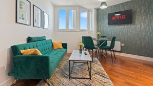 A modern living room with a green sofa, a coffee table, and a dining area with chairs. A television is mounted on the wall, and there are framed pictures on the wall. - Emerald Suite- Bham Citycentre- Luxury-cinema Room (Birmingham)