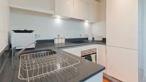 A modern kitchen with white cabinets, a black countertop, a kettle, a toaster, and a sink with a drying rack. - Emerald Suite- Bham Citycentre- Luxury-cinema Room (Birmingham)
