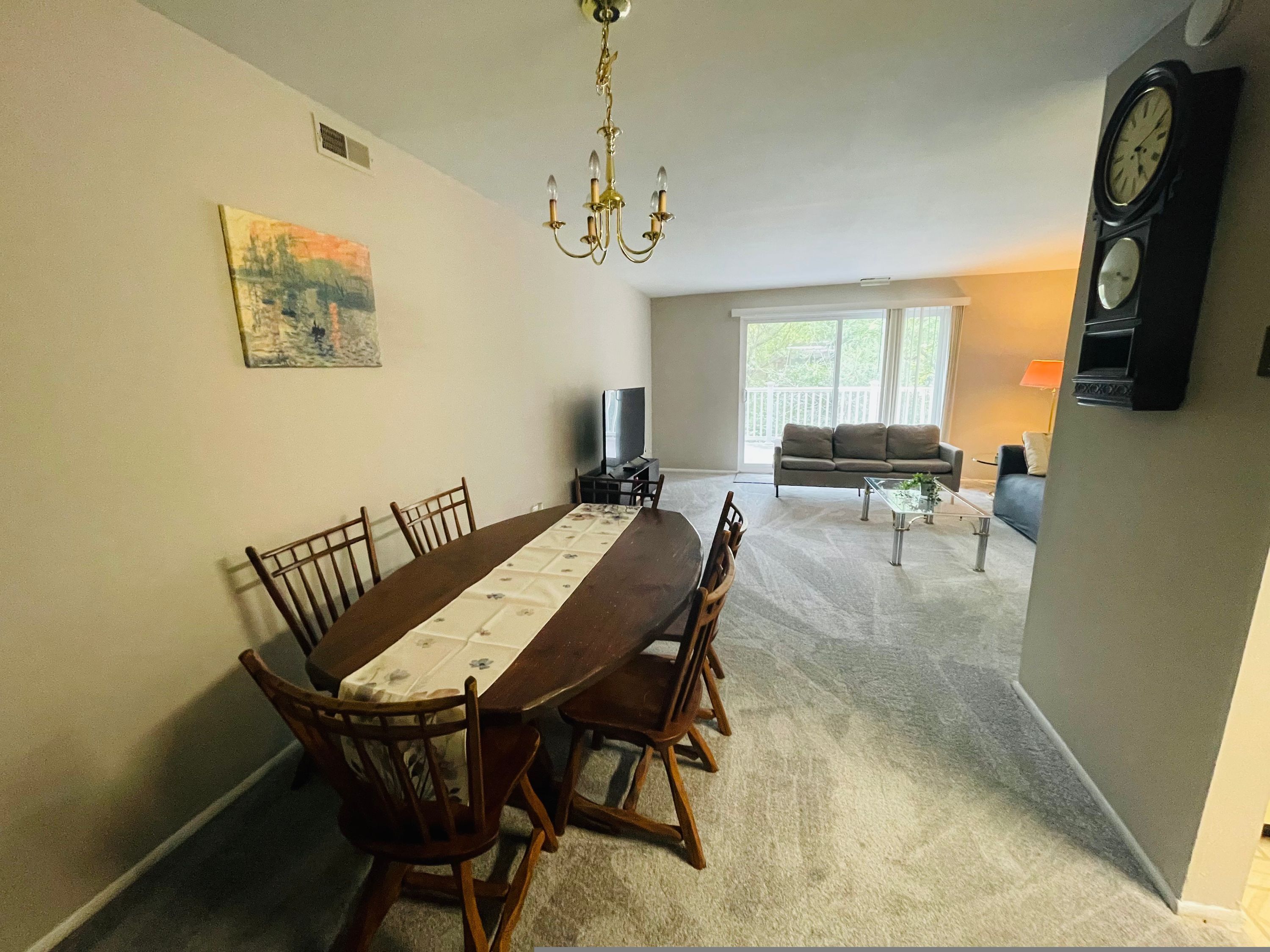 A dining area with a long table, chairs, a chandelier, a painting, a sofa, a TV, and a clock.
