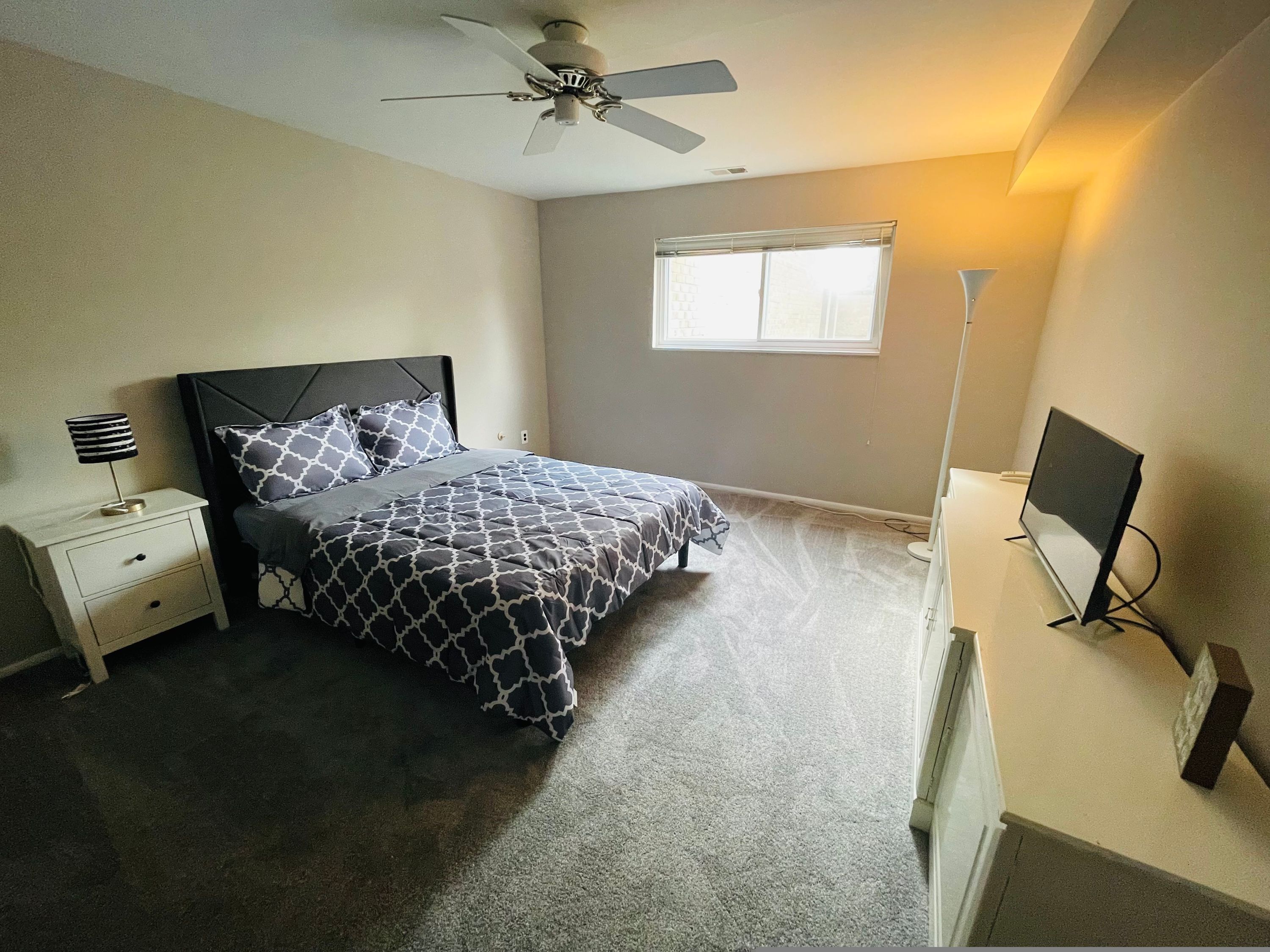 A bedroom with a bed, a nightstand, a ceiling fan, a window, and a TV stand.