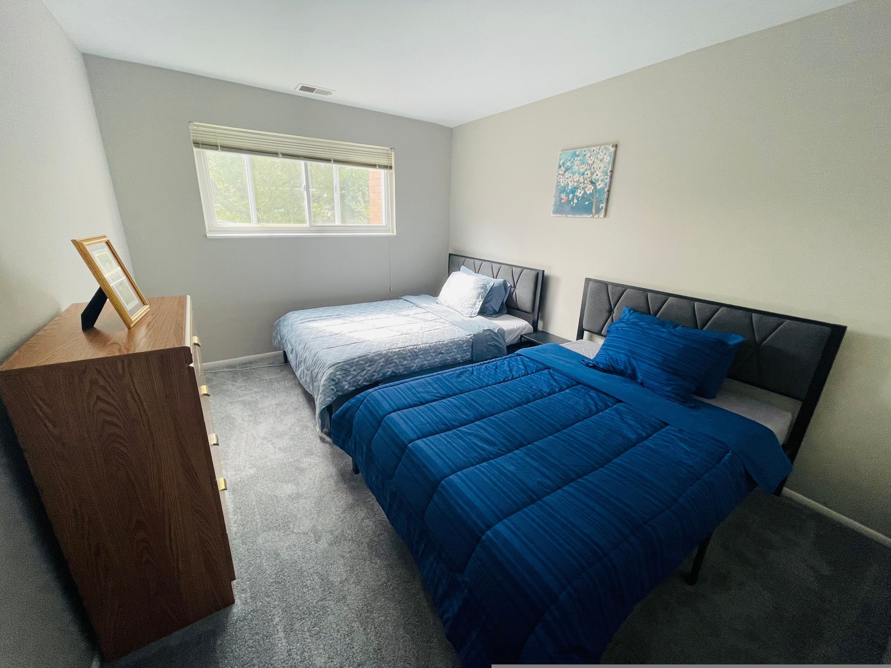 A bedroom with two beds, a wooden dresser, a window with blinds, and a painting on the wall.