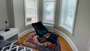 A corner bedroom with three large windows, a radiator, a dark blue chair, a patterned rug, and a white bedside table. - Charming Place to Call Home (Elkins Park)