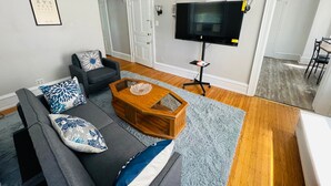 A living room with a grey sofa, a wooden coffee table, a flat-screen TV on a stand, and a door leading to another room. - Charming Place to Call Home (Elkins Park)