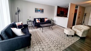 Ein modernes Wohnzimmer mit einem dunkelblauen Sofa, einem gemusterten Teppich und einem Couchtisch. An der Wand hängt ein Gemälde, und durch eine offene Tür ist eine Küche erkennbar.