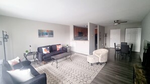 Ein geräumiges Wohnzimmer mit einem dunkelgrauen Sofa, einem gemusterten Teppich und einem Essbereich mit Tisch und Stühlen.