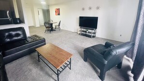 Un salon avec un canapé en cuir noir, une table basse en bois, un téléviseur posé sur un meuble et un coin repas comprenant des chaises et une table.