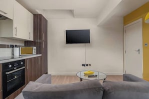 A modern kitchen with a microwave, oven, and cabinets, a grey sofa, a small glass table with a yellow object, and a TV mounted on the wall.