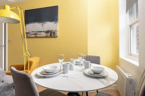 A dining area with a round table set for four, a yellow floor lamp, a painting on the wall, and a window on the right.