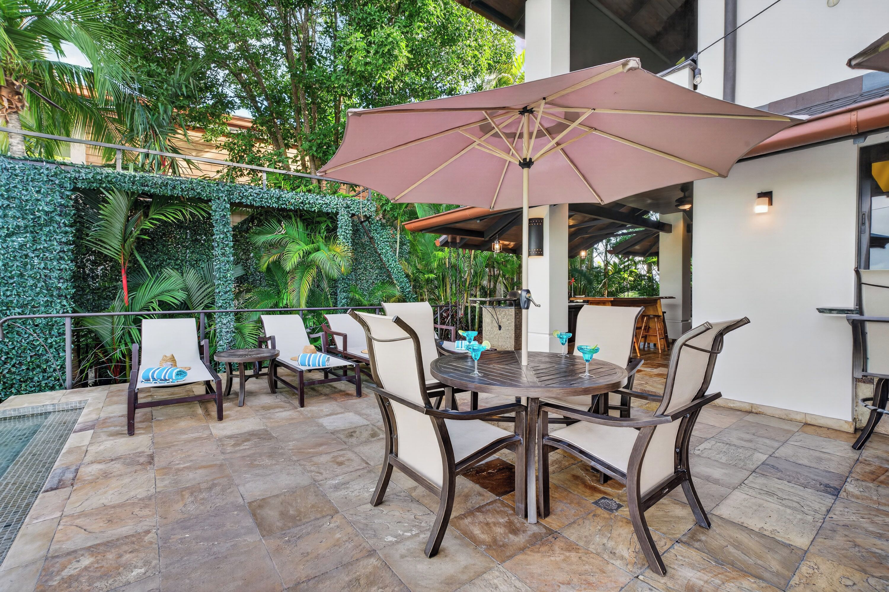 An outdoor patio with a table and chairs, surrounded by lush greenery and a covered area with an umbrella.