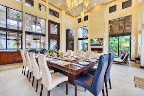 A spacious dining area with a long table set for a meal, surrounded by chairs, and large windows offering views of the outdoors.