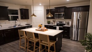 Una cocina moderna con armarios oscuros, electrodomésticos de acero inoxidable, una isla central con sillas e iluminación colgante.