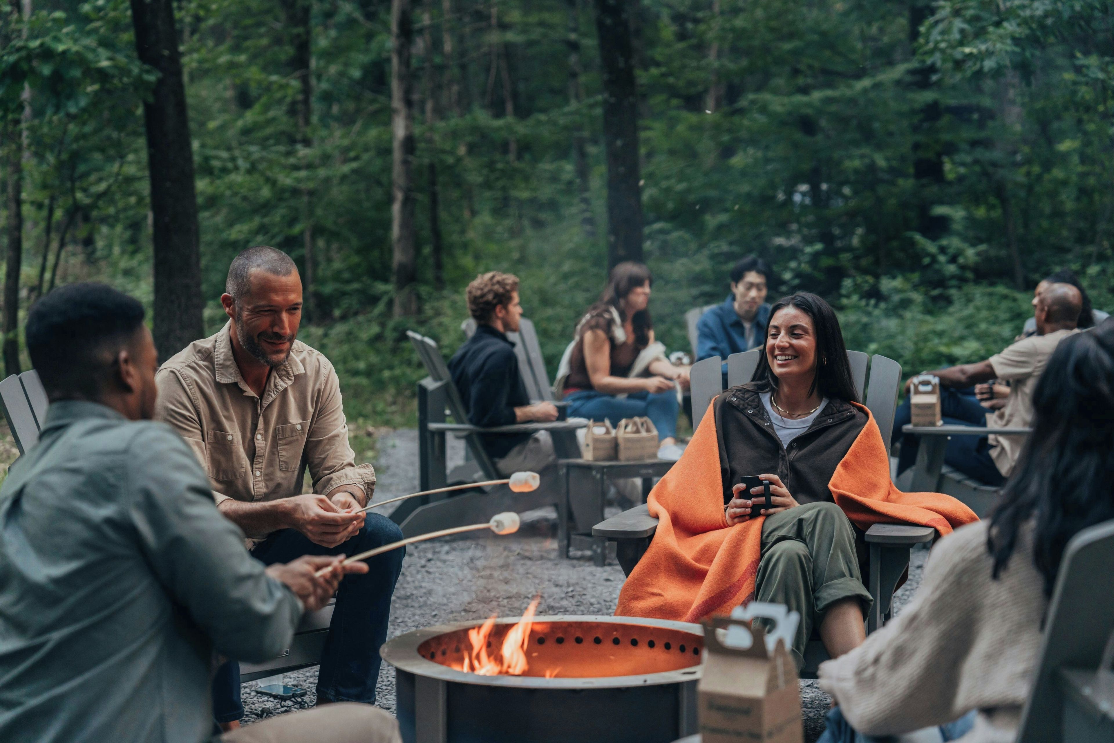 Un grupo de personas reunidas alrededor de una fogata en un entorno natural, con árboles al fondo.