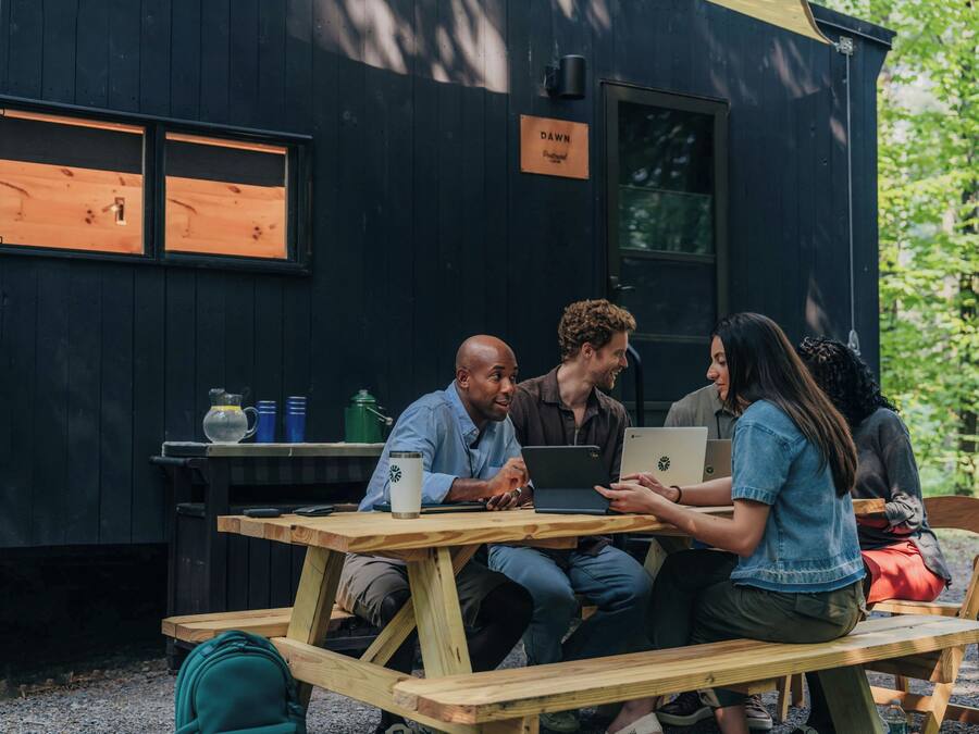 A group of people sitting at a wooden picnic table outside a black building with a sign that reads 'DAWN'.