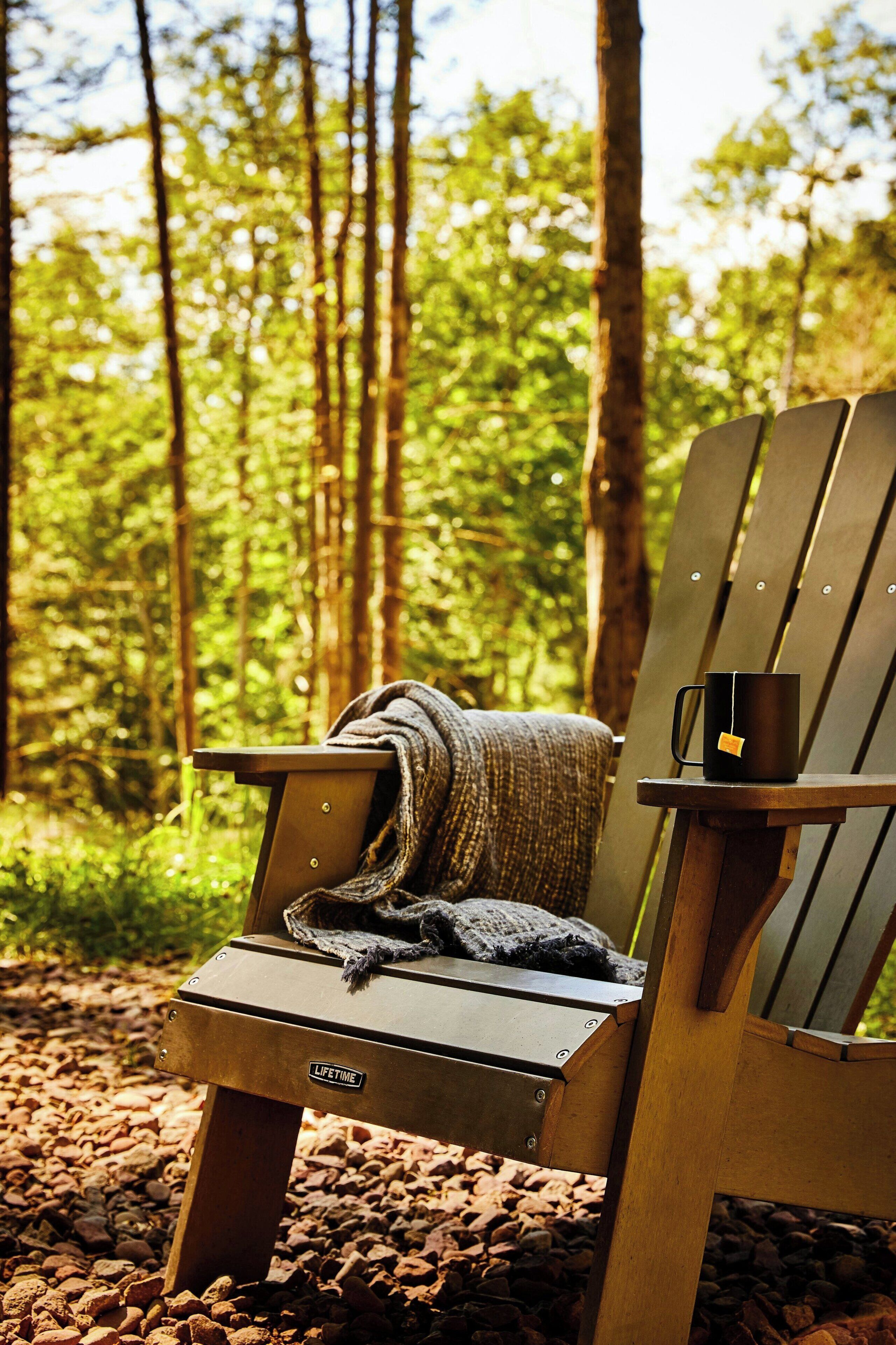 A wooden Adirondack chair with a blanket and a mug in a forest setting.