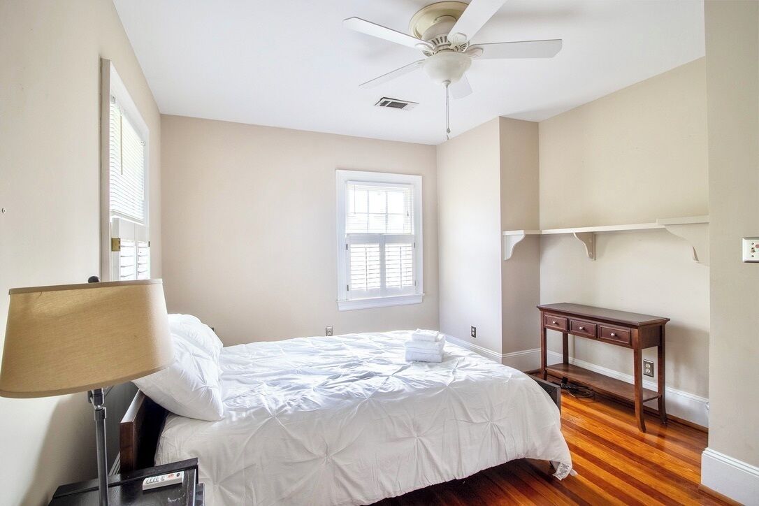 Une chambre à coucher comprenant un lit, une table de chevet, une lampe, un ventilateur de plafond, une fenêtre et une petite table en bois.