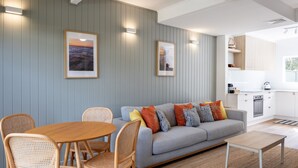 A modern living room with a grey sofa, colorful cushions, wicker chairs, and a wooden coffee table. There are framed pictures on the wall and a kitchen area visible in the background.