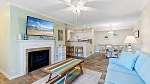A modern living room with a fireplace, a television, a dining area, and a kitchen. - Colony Club H6 with Pool (Gulf Shores)