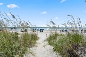 En sti af sand der fører hen til havet, med strandparasoller og liggestole.