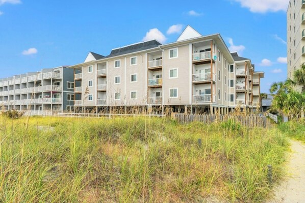 A multi-story hotel with balconies, located near a beach with grassy dunes.