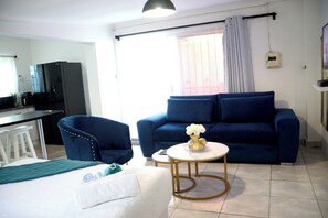 Relax in this stylish living room with a sofa, armchair, and coffee table, featuring a kitchen area and large window with a curtain.