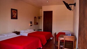 A room with two beds, each with red bedding, a wooden chair, a small table, a wall-mounted lamp, and a door.