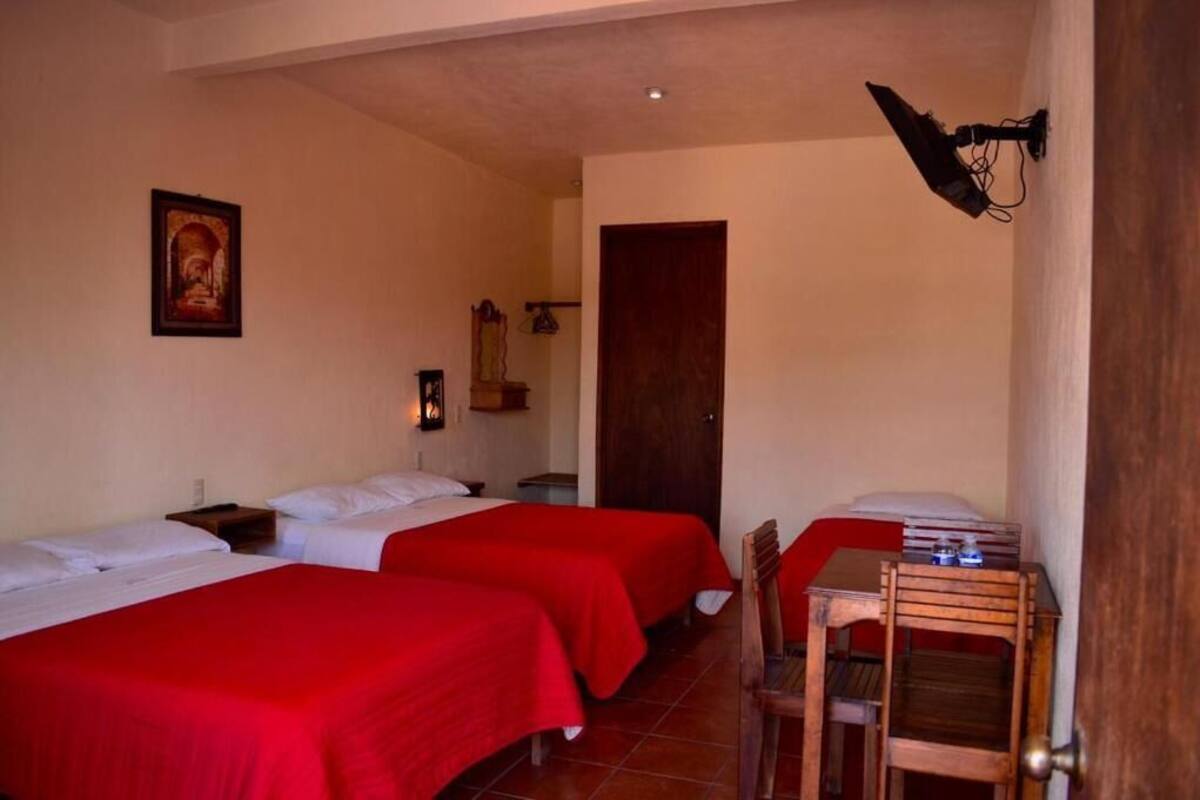 A room with two beds, each with red bedding, a wooden chair, a small table, a wall-mounted lamp, and a door.