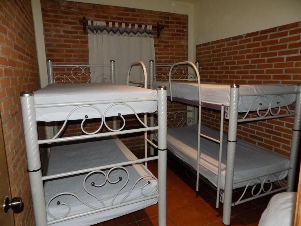 A room with three bunk beds against a brick wall, a window with white curtains, and a door with a metal handle.