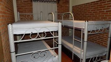 Una habitación con tres literas contra una pared de ladrillo, una ventana con cortinas blancas y una puerta con manija de metal.