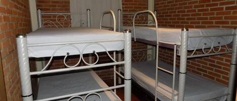 Una habitación con tres literas contra una pared de ladrillo, una ventana con cortinas blancas y una puerta con manija de metal.