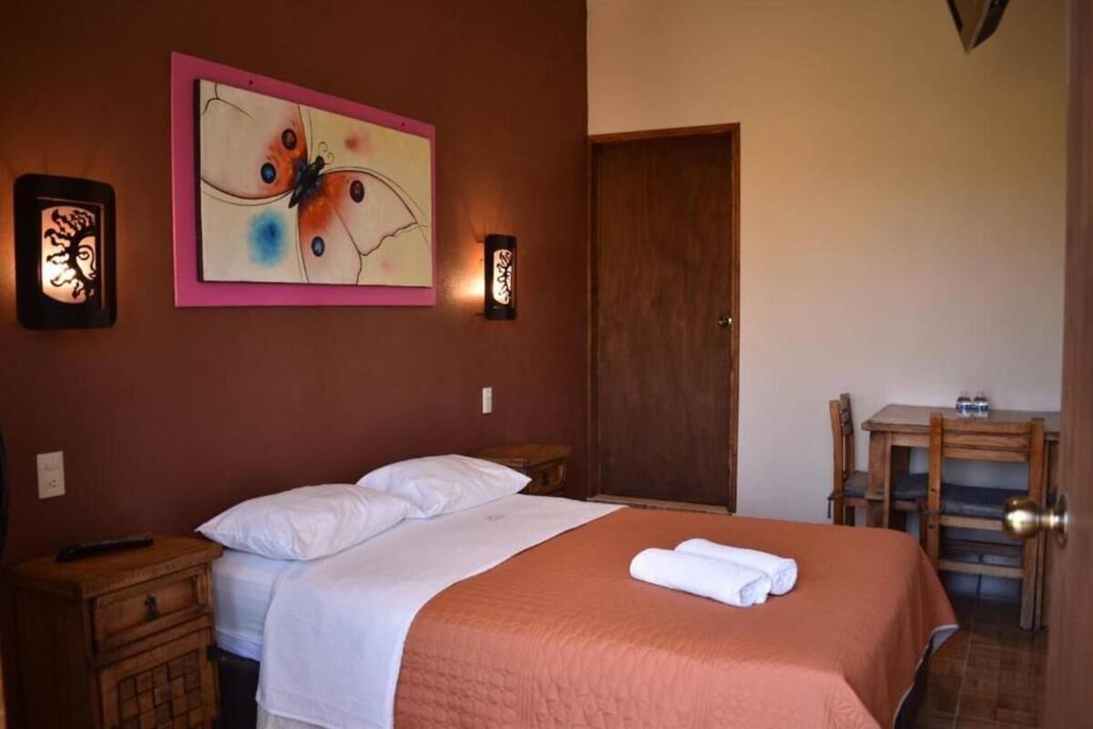 A bedroom with a bed, a nightstand, a bedside table, a wooden chair, a small table with a bottle, and a door.