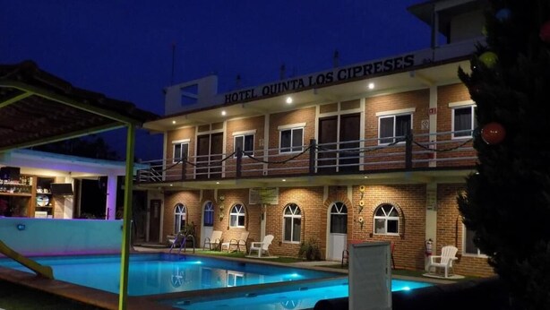A hotel with a swimming pool, outdoor seating, and a sign reading "HOTEL QUINTA LOS CIPRESES".