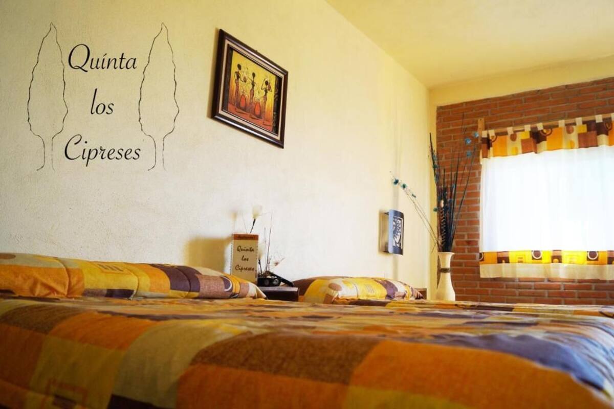 A hotel room with a bed, a framed picture on the wall, a small table with a vase, and a window with curtains.
