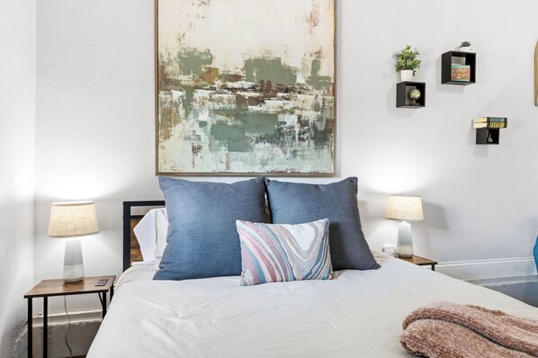 A neatly made bed with two pillows, a bedside table with a lamp, a wall-mounted abstract painting, and a small shelf with decorative items. - Near HSU, ACU & Hendrick ~ Clean ~ Charming Cottage ~ New Discounts ~ Dogs (Abilene)