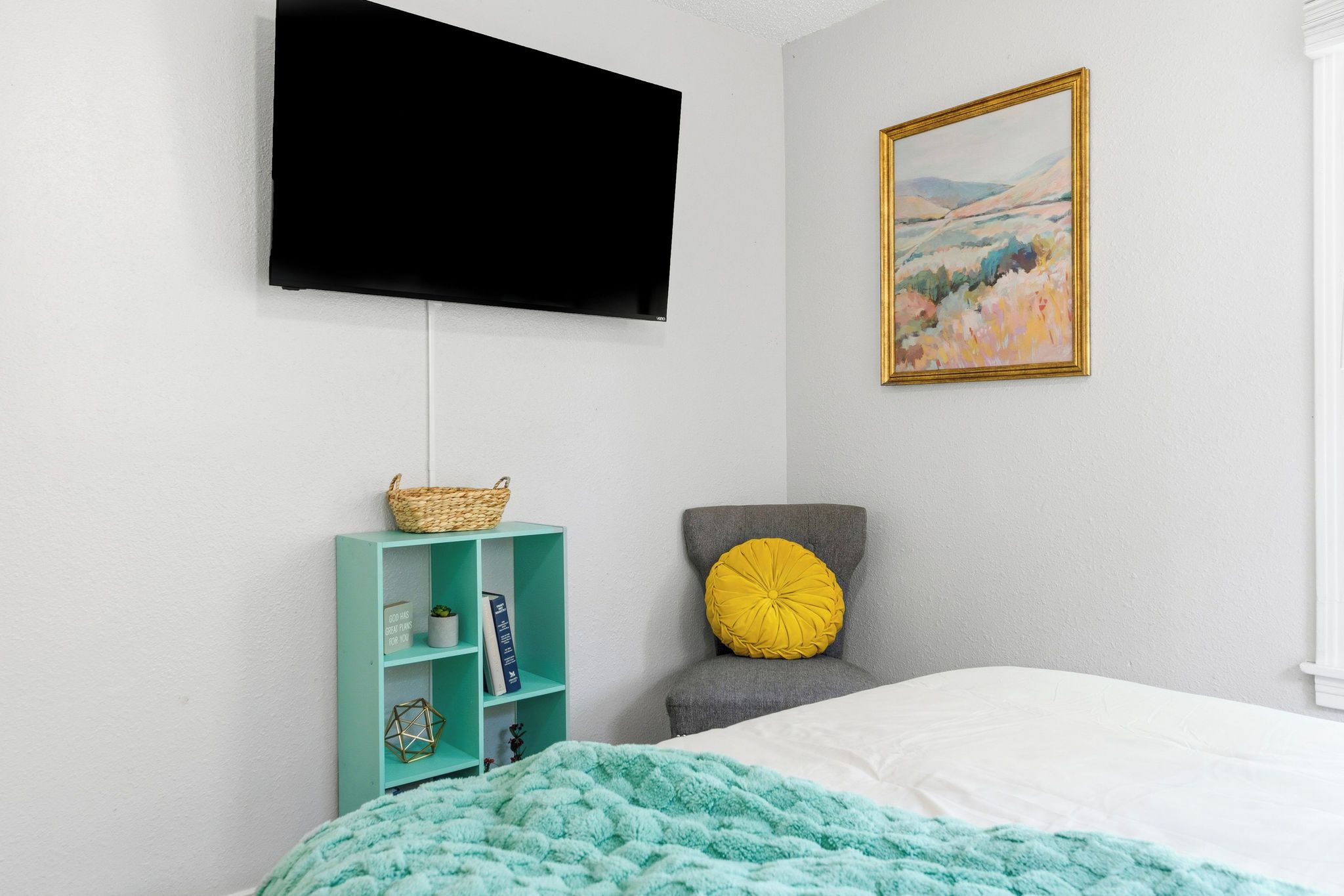 Un dormitorio con cama, una estantería verde azulada, un sillón gris con almohada amarilla, un televisor montado en la pared y un cuadro de paisaje.
