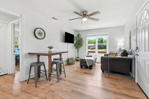 A spacious room with a ceiling fan, a dining table with stools, a sofa, a potted plant, and a television mounted on the wall. - Near HSU, ACU & Hendrick ~ Clean ~ Charming Cottage ~ New Discounts ~ Dogs (Abilene)