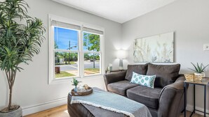 A living room with a brown sofa, a potted plant, a window with blinds, and a wall-mounted lamp. - Near HSU, ACU & Hendrick ~ Clean ~ Charming Cottage  ~ New Discounts ~ Dogs (Abilene)