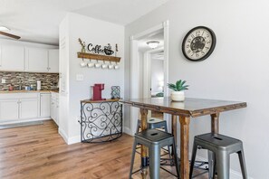 A kitchen with white cabinets, a coffee-themed wall decoration, a clock, and a wooden table with stools. - Near HSU, ACU & Hendrick ~ Clean ~ Charming Cottage ~ New Discounts ~ Dogs (Abilene)