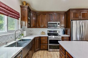A kitchen with wooden cabinets, stainless steel appliances, and a window with a red valance. - Timp Retreat | EV | Free Massage* | Backyard (Orem)