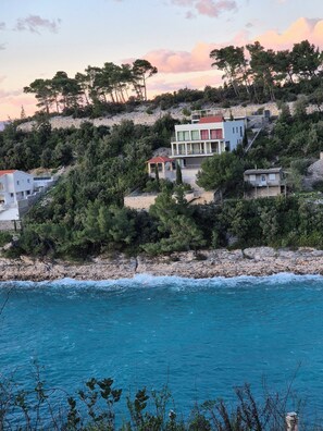 Il s’agit d’une propriété située sur la côte, comprenant plusieurs bâtiments, notamment une maison à deux étages avec un toit rouge et un bâtiment blanc doté d’un balcon. La propriété est entourée d’une végétation luxuriante et d’un terrain rocheux.