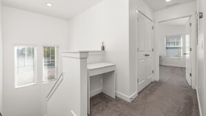 A hallway with white walls, a carpeted floor, a small white desk, and a door leading to another room.