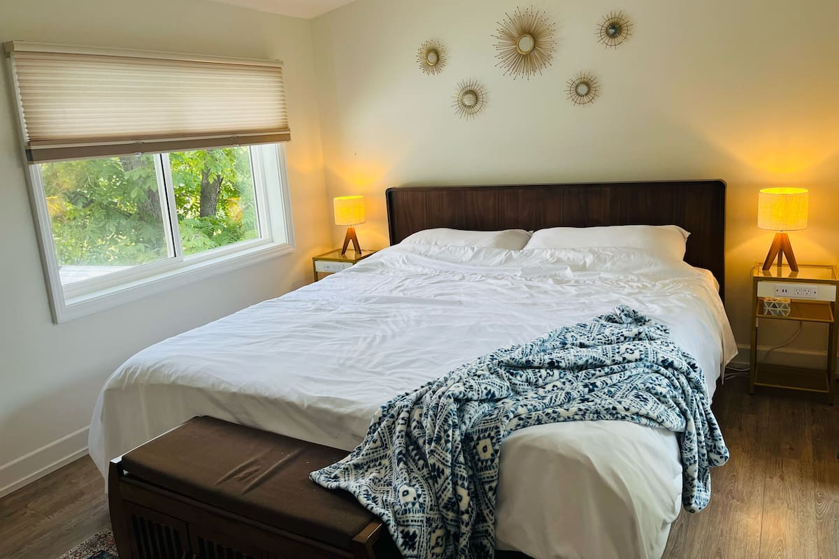 A neatly made bed with a patterned blanket, two bedside lamps, a window with blinds, and wall decor.
